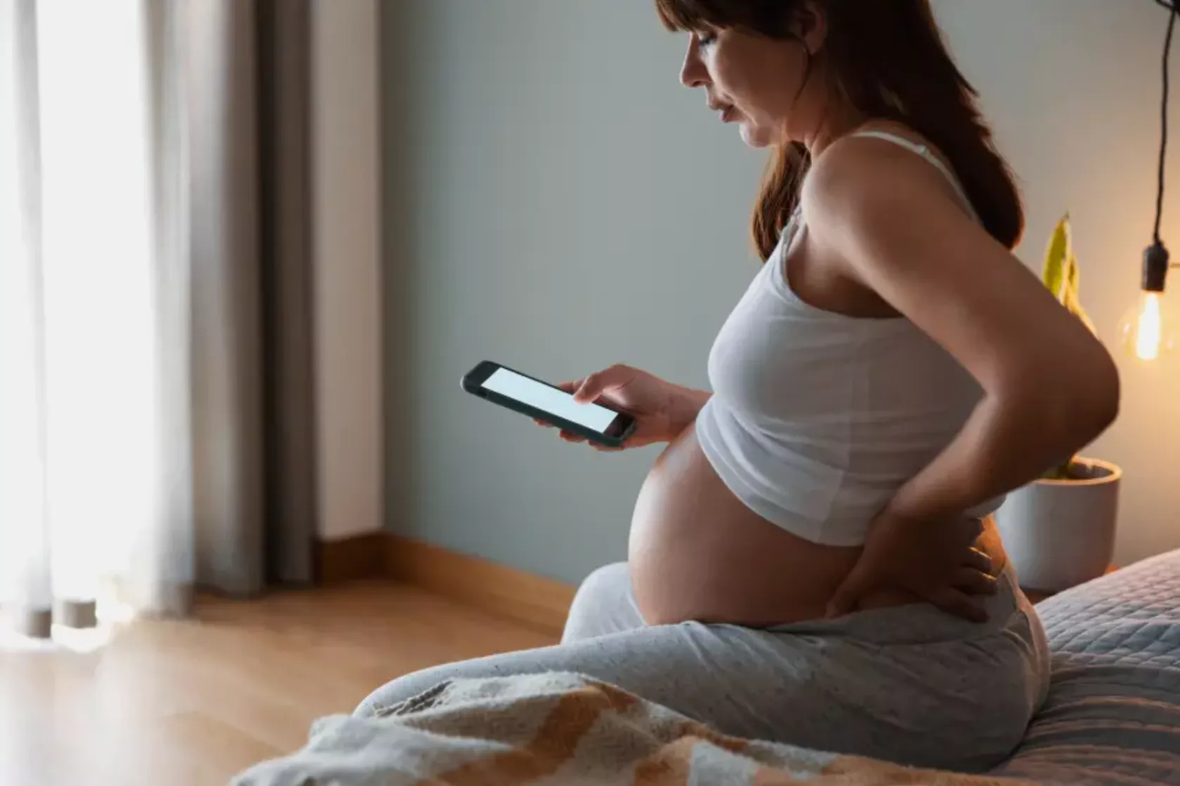 Schwangere Frau sitzt mit Handy in der Hand auf dem Bett