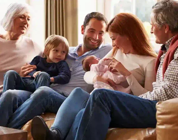 Paar mit Neugeborenem, älterem Kind und Großeltern fröhlich auf dem Sofa.