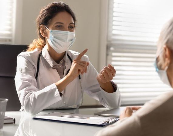 Ärztin lächelt hinter ihrem Mundschutz und unterstreicht ihre Wort im Beratungsgespräch mit Hand-Gesten.