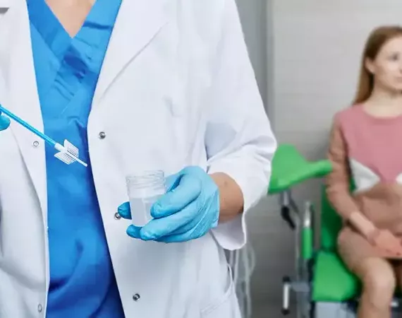 Cyto-Brush in der Hand des Gynäkologen beim PAP-Abstrich, im Hintergrund sitzt die Patientin.