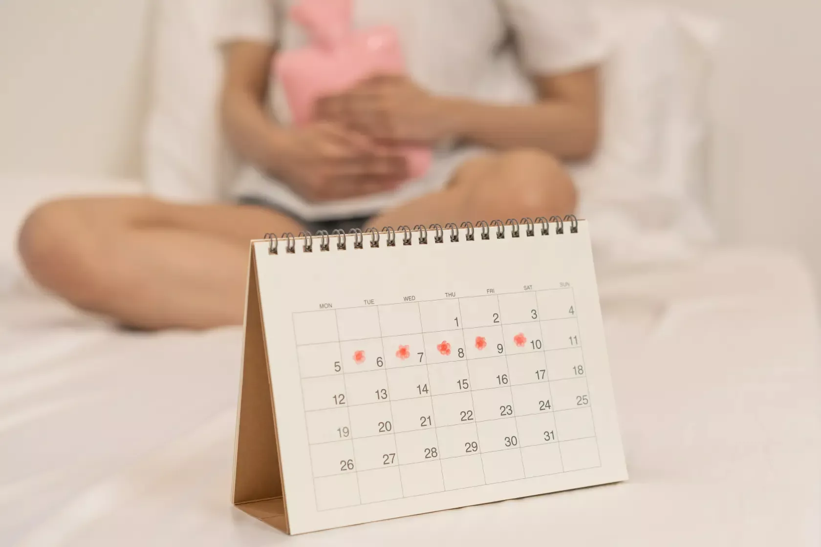 Frau sitzt mit Wärmflasche auf dem Bett. Vor ihr der Menstruationskalender.