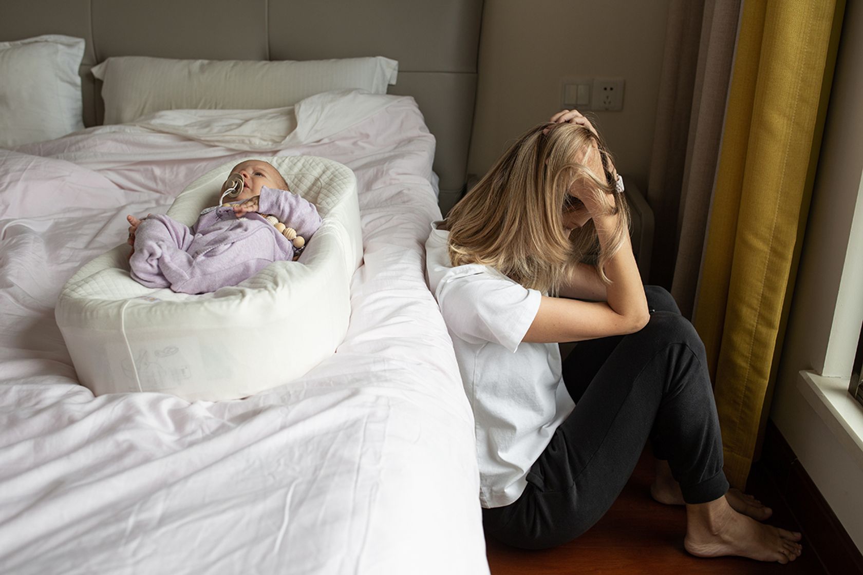 Mutter sitzt neben dem Bett, auf dem ihr Säugling liegt. Sie stützt mit verzweifeltem Ausdruck den Kopf in die Hände.