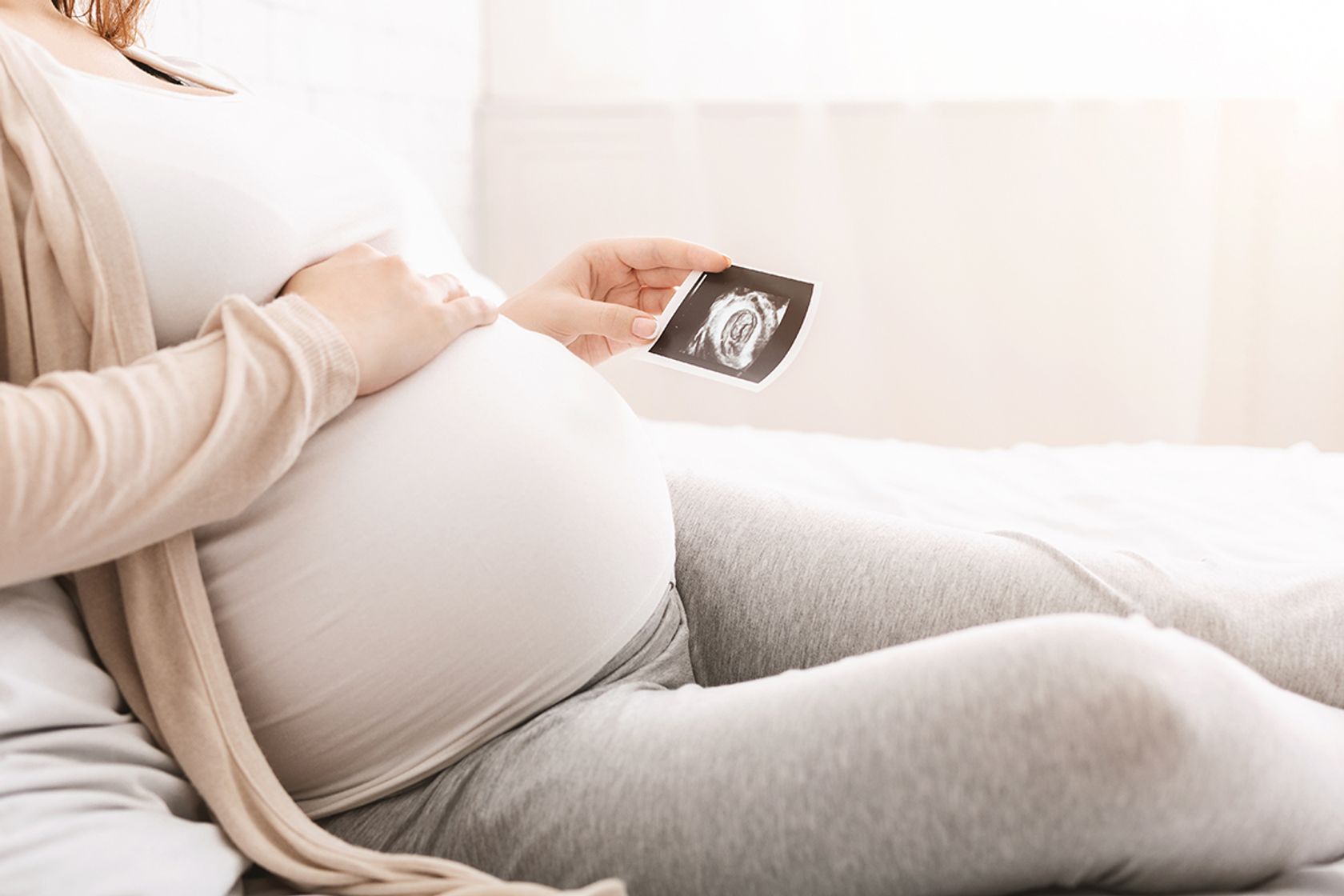 Eine Schwangere hält ein Ultraschallfoto ihres Babys in der Hand und ihre andere Hand liegt auf ihrem Bauch.