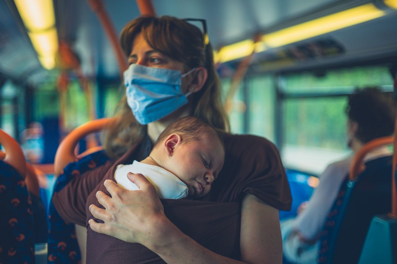 Junge Frau mit Mundschutz hält ihr Baby im Arm in den öffentlichen Verkehrsmitteln