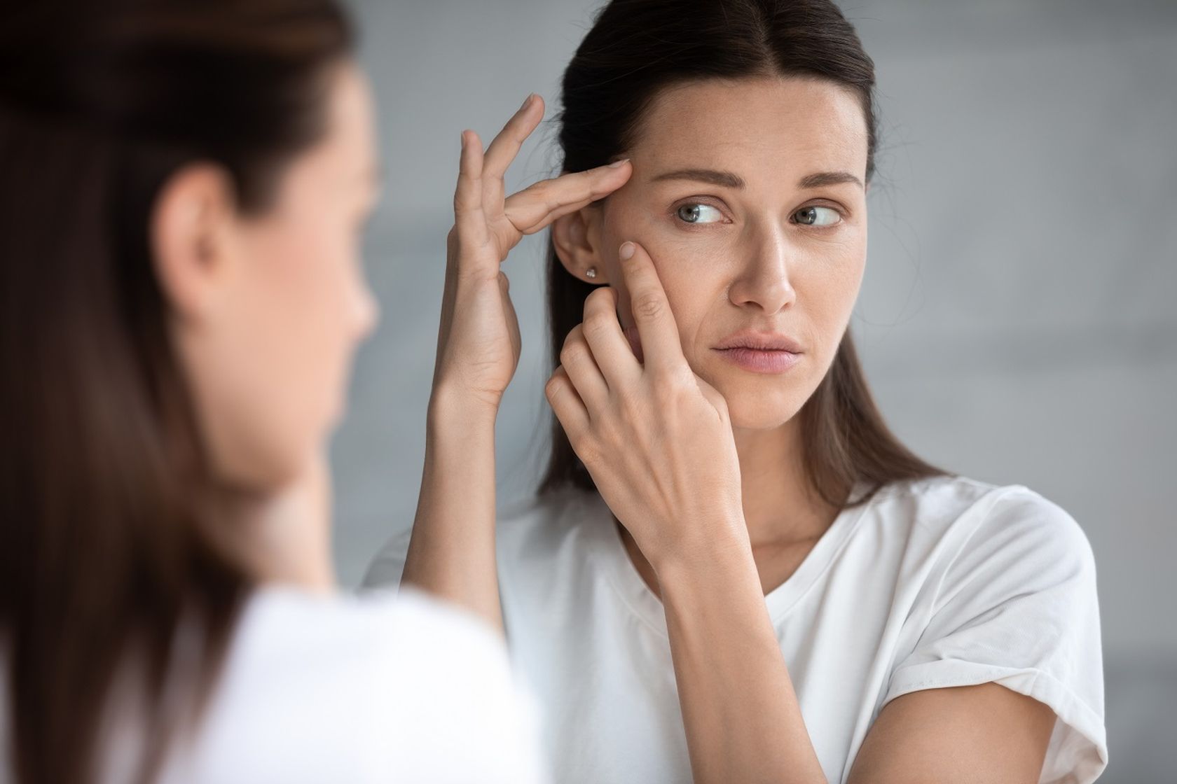 junge Frau betrachtet mit sorgenvoller Miene die Haut in ihrem Gesicht im Spiegel