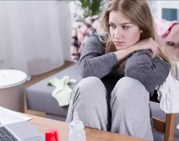 Junge Frau mit deprimiertem Gesichtsausdruck sitzt vor einem Tisch mit Babyfläschchen und Laptop.