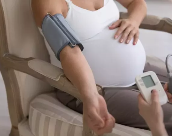Close-up einer hochschwangeren Frau, die auf einem Stuhl sitzt und bei der mit Manschette am Arm der Blutdruck gemessen wird.