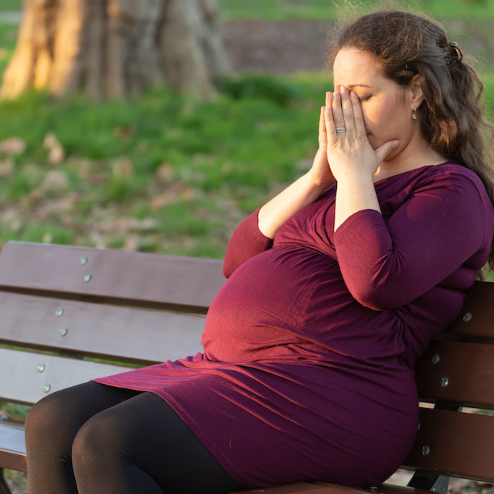 Schwangere auf einer Parkbank sitzend hält ihre Hände vors Gesicht.