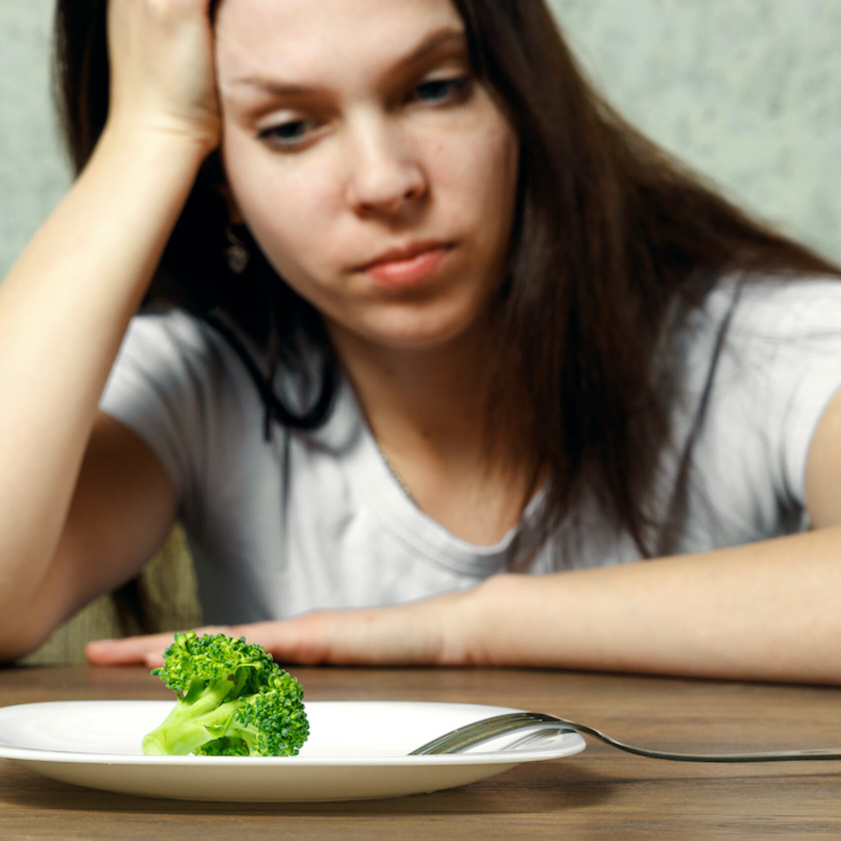 Junge Frau sitzt appetitlos vor einem Teller mit ganz wenig Gemüse und kann oder möchte nicht essen.
